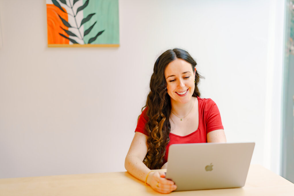 Image of a white women in her 30s with long curly brunette hair smiling warmly at a laptop while holding the sides