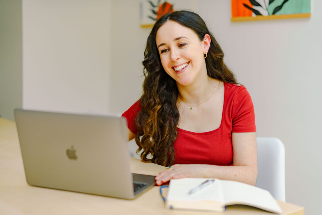 Image of a white women in her 30s wearing a burgundy t-shirt smiling warmly at a laptop